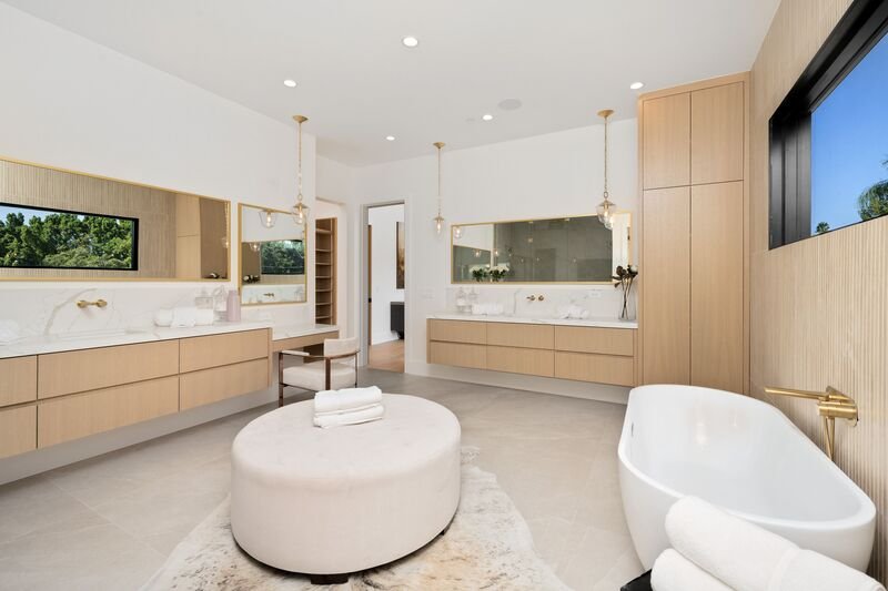 nterior de baño moderno y espacioso con bañera exenta, lavabo doble y espejos grandes. La luz natural que entra por las ventanas ilumina el espacio